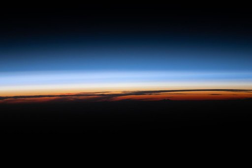 A sunset below Earth’s horizon silhouettes the clouds and illuminates the atmosphere