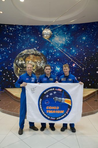 During a break from training, the Expedition 46-47 crewmembers toured the Korolev Museum at the Baikonur Cosmodrome in Kazakhstan Dec. 10 and displayed a flag bearing the logo for their Soyuz mission. From left to right are Tim Kopra of NASA,  Yuri Malenchenko of the Russian Federal Space Agency (Ro