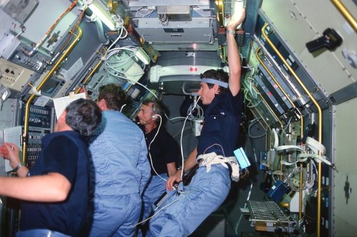STS-9 crewmembers gather around television monitor in Spacelab module