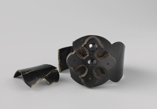 Bracelet of whalebone, broad at the front and somewhat wavy. On top a Greek cross; decorated with studs