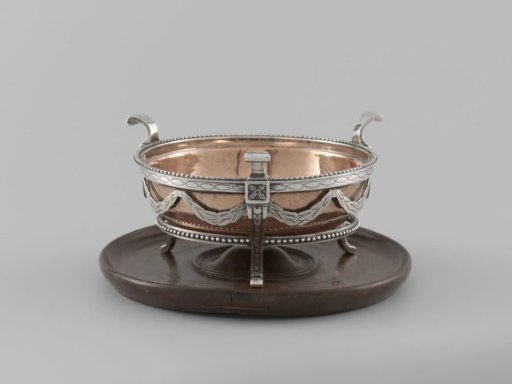 Tobacco comforter on a wooden board. The inner copper bowl hangs in an open silver frame resting on the board with three feet.