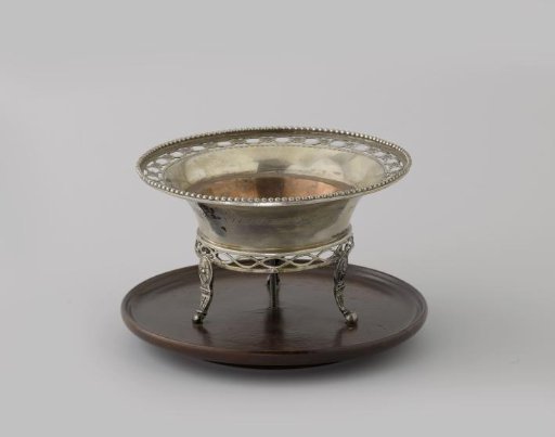 Silver pipe warmer on a wooden tray. Round vessel on three legs with a flared rim. Decorated along the lower edge with a pierced scroll with rosettes.