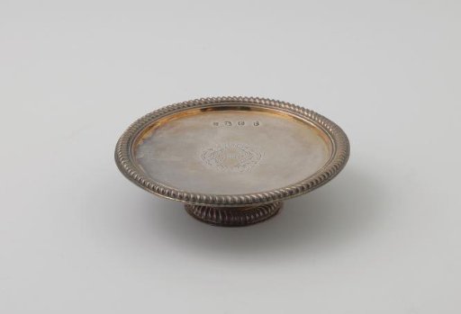 Silver presentation plate, round, edged along the rim with a heavy cable border. On the field a lozenge-shaped coat of arms between leaf and cartouche motifs. Short, round foot.