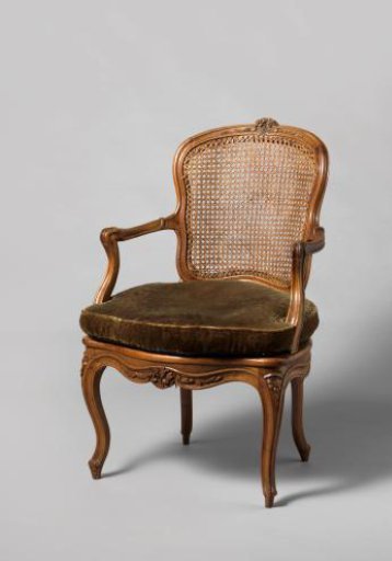Armchair of walnut with S-shaped splayed legs with carving of flowers and leaves. Back and seat of woven reed. With loose cushion.