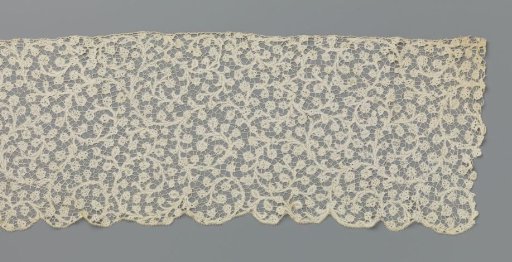 Strip of bobbin lace with volute scrolls and round flowers on curling side branches