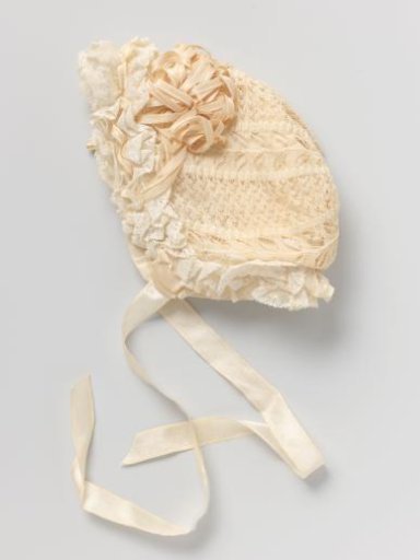 Children's cap of openwork knitted cream-colored cotton, decorated with loops and two rosettes of narrow, cream-colored satin ribbon.