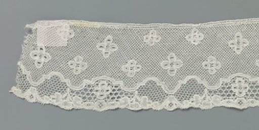 Strip of bobbin lace with a symmetrical pattern around a continuous scalloped decorative border below the center line of the strip