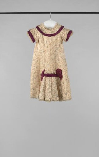 A party dress of printed cream silk with flower sprigs, trimmed with carmine red ribbons and strips. The apron is of glass batiste, and under the skirt hem Valenciennes lace appears
