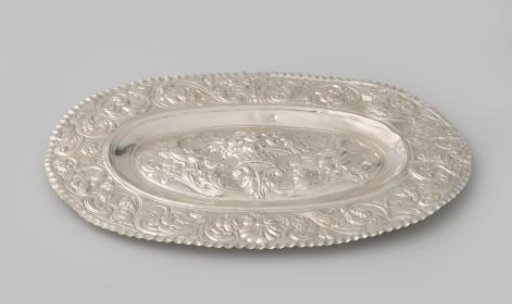 Silver leaf, oval with a broad flat rim with chased floral and foliate scrolls. On the slightly domed surface a fruit basket flanked by two birds.