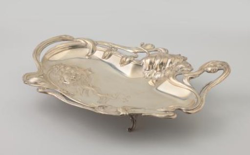 Silver salver, oblong and irregularly shaped, with a rim decorated with flower stems and leaves. On the flat surface a woman's head with wavy hair merging into the rim.