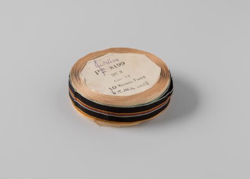 Ribbon on a roll, striped, the ribbon is black with three woven stripes in the center in the colors orange, red and light blue, and is edged on the sides with light blue and orange embroidery respectively