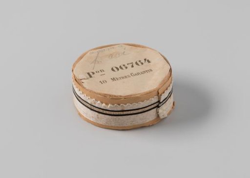 Ribbon on a roll, white satin with a central stripe of openwork flanked by two black stripes, one side scalloped and edged with a wavy white border