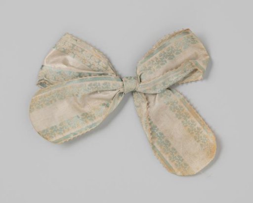 Bow of cream silk, decorated along the edges with a stripe and a leaf vine embroidered in blue silk