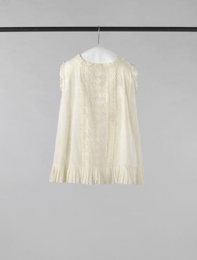 Sleeveless child's pinafore of white cotton with a wide central band of English embroidery; pleated at the front bottom and trimmed with English embroidery along the neckline and armholes.