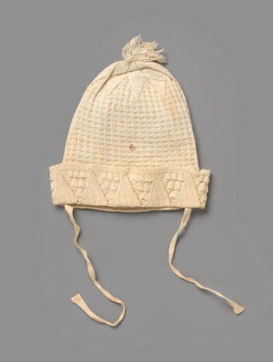Knitted cap of white cotton with vertical ribbed borders, between which a decorative motif, flower shape, tassel and turned-down scalloped border with triangles with decorative motif