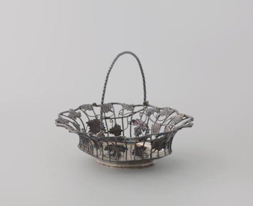 Silver basket handle. Composed of curved rods rising from the base and connected at the top by a wavy rim. Decorated with winding grapevines.