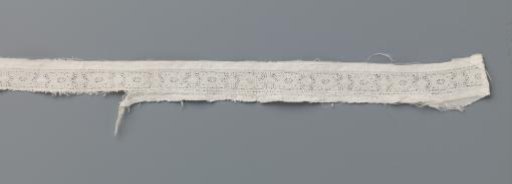 Small strip of bobbin lace with stylized flower between two vertical lines and ovals on a rectangular fragment of batiste
