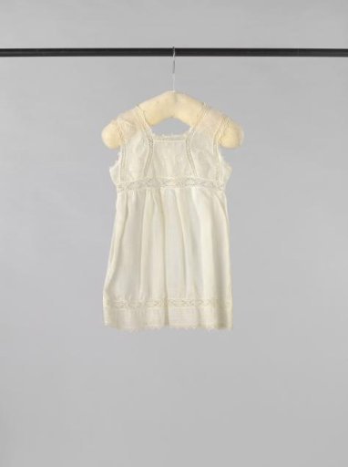 Sleeveless pinafore of white cotton (?), decorated with openwork borders and on the bodice with white embroidered purl stitches and twice a stylized flower and leaf motif