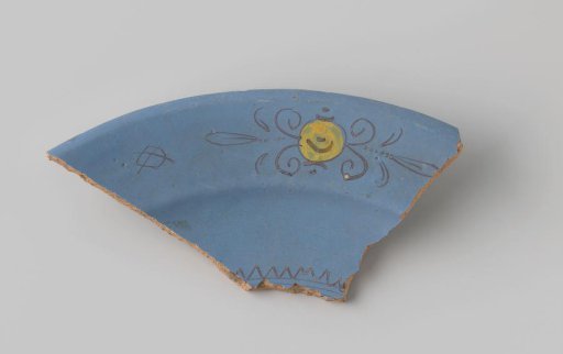 Fragment of reddish-fired earthenware, glazed blue on the front, with a diamond, a scalloped edge and a stylized flower form filled with yellow painted in brown
