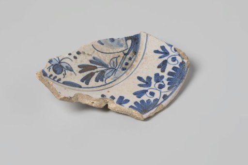 Sherd of a plate of earthenware with white tin glaze on the back and tin glaze on the front and a decoration of flowers and insects in blue, damaged