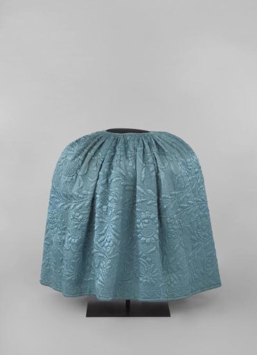 Skirt of blue silk, quilted and stitched with very large floral and foliate scrolls to the top; the strongly pleated skirt has a brown linen lining.
