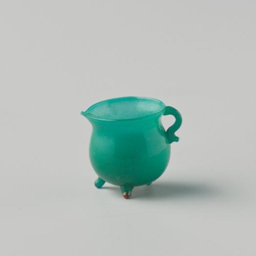 Cooking jug of opaque green glass (probably imitation green earthenware); on three feet, with spout and handle.