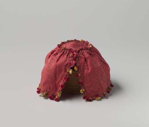 Fire basket cover of red silk with a woven diamond pattern, trimmed with passementerie with yellow and red tassels