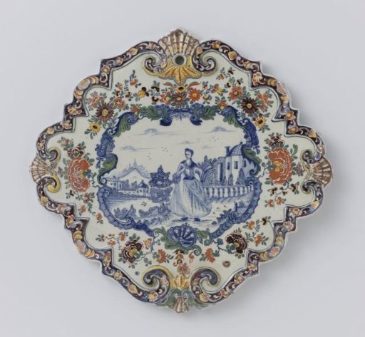 Plate, diamond-shaped with scalloped edge. Within multicoloured flower border a scene in blue painted of a lady in a garden