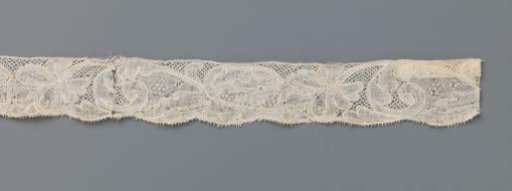 Strip of bobbin lace with a palm-shaped flower on a long curved stem with a volute leaf