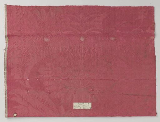 Fragment of red silk damask with a pattern of stylized flower with vase-shaped ovary and petals, surrounded by curved leaves and garlands
