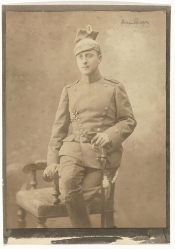 Arthur Moos with a chapka and sabre in the uniform of the German Army, with the Iron Cross on his chest, seated on a chair back, December 1914