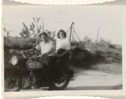 Leo Blumensohn and Isabel Wachenheimer seated on a motorcycle, standing in front of a fence and a vegetated slope, 1945–1949