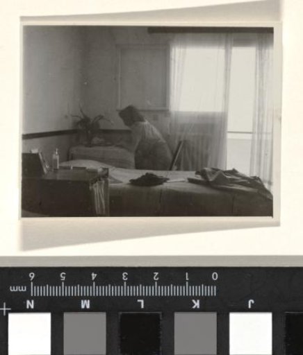 Isabel Wachenheimer seated before an open window with curtains before a bed, writing at a table