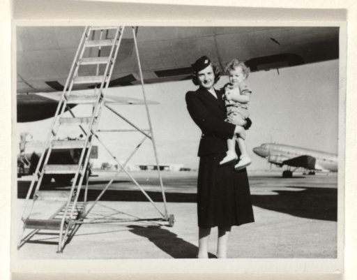 Isabel Wachenheimer in KLM uniform, standing next to a staircase and in front of an airplane, with a child in her arms