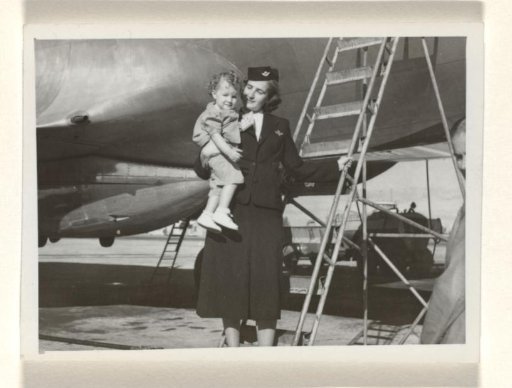 Isabel Wachenheimer in KLM uniform, standing before a staircase and in front of an airplane, with a child in her arms