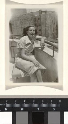 Isabel Wachenheimer in a dress with a pattern of striped bands, seated on a chair with her arm on the railing of a balcony
