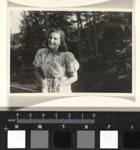 Isabel Wachenheimer in a dress with a floral pattern and her hands in her pockets, standing in front of a forest