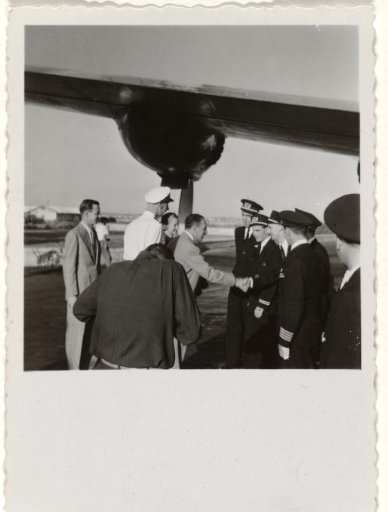 Ground staff greeting a man in a dark suit in front of an airplane, with a photographer in the foreground