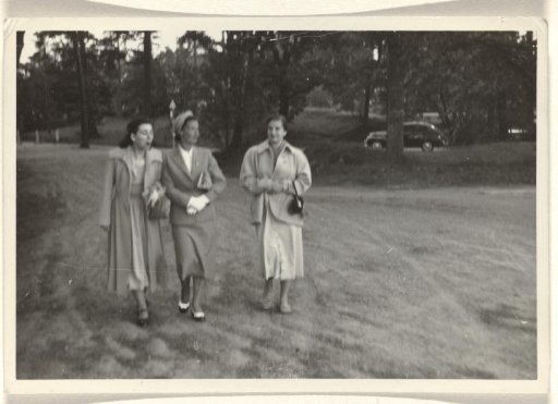 Isabel Wachenheimer in a long coat and two unknown women, walking across a grassy field with trees and a car in the background