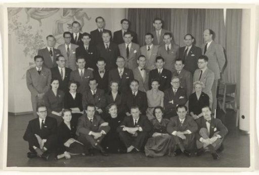 Group portrait of 37 unknown people and Isabel Wachenheimer, in front of a patterned wall and drawn curtains