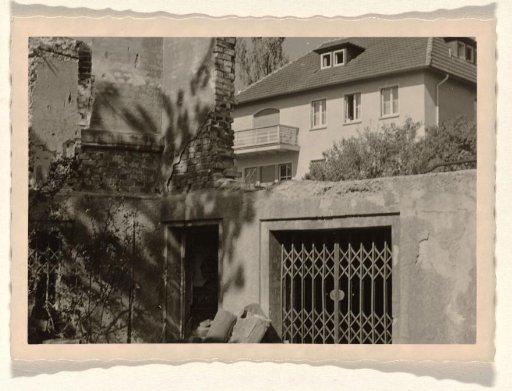 Part of a ruin with a sliding gate and a door opening in it, with a large house with a balcony in the background, surrounded by trees and shrubs