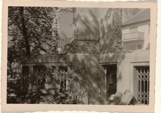 Part of a ruin with a sliding gate and a doorway in it, with a large house with a balcony in the background, with trees and bushes around it