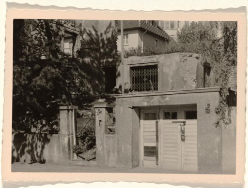 Part of a ruin with a sliding gate and a doorway in it, with a large house with a balcony in the background, with trees and bushes around it