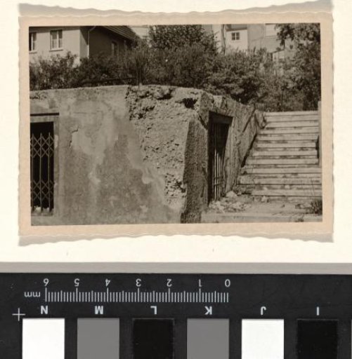 Part of a ruin with a sliding gate and a doorway in it, with a large house with a balcony in the background, with trees and bushes around it