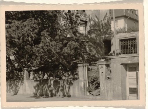 Part of a ruin with a sliding gate and a doorway in it, with a large house with a balcony in the background, with trees and bushes around it