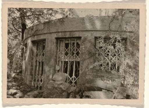 Circular part of a ruin, with bars on the windows, stones in the foreground and vegetation on the right
