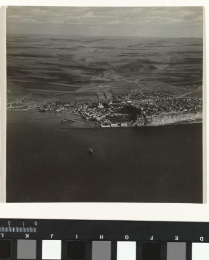 Black-and-white aerial photograph of a town or village surrounded by meadows, situated on the water