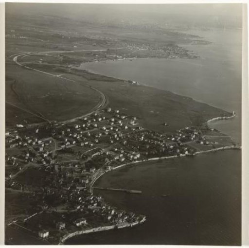Aerial photograph in black and white of a town or village situated on the water, with meadows in the background, located on a bay with a quay