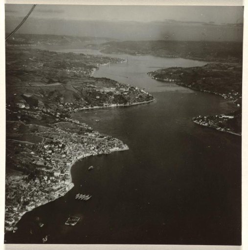 Aerial photograph in black and white of a town or village situated on a river or bay, with inlets in the river or bay and boats and meadows