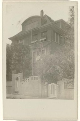 Postcard of a large building with open windows on the left side of the building, and a fence and vegetation in the foreground